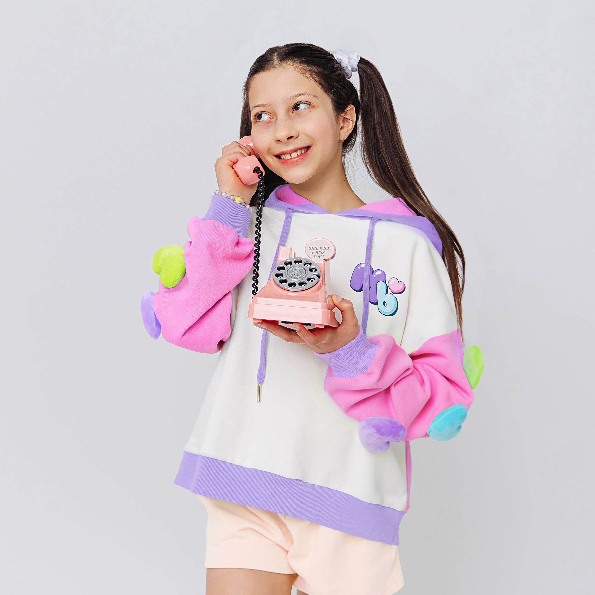 A girl wearing a cool colorful hoodie with rainbow 3D hearts plush accessories attached on sleeves holding a toy phone.