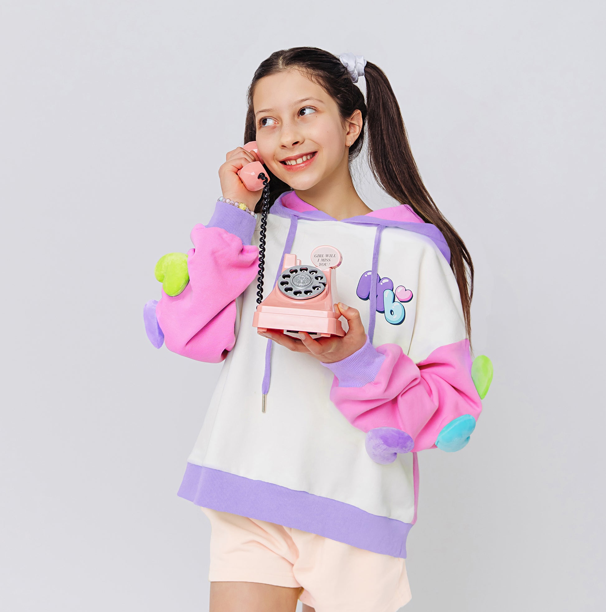 A girl wearing a cool colorful hoodie with rainbow 3D hearts plush accessories attached on sleeves holding a toy phone.