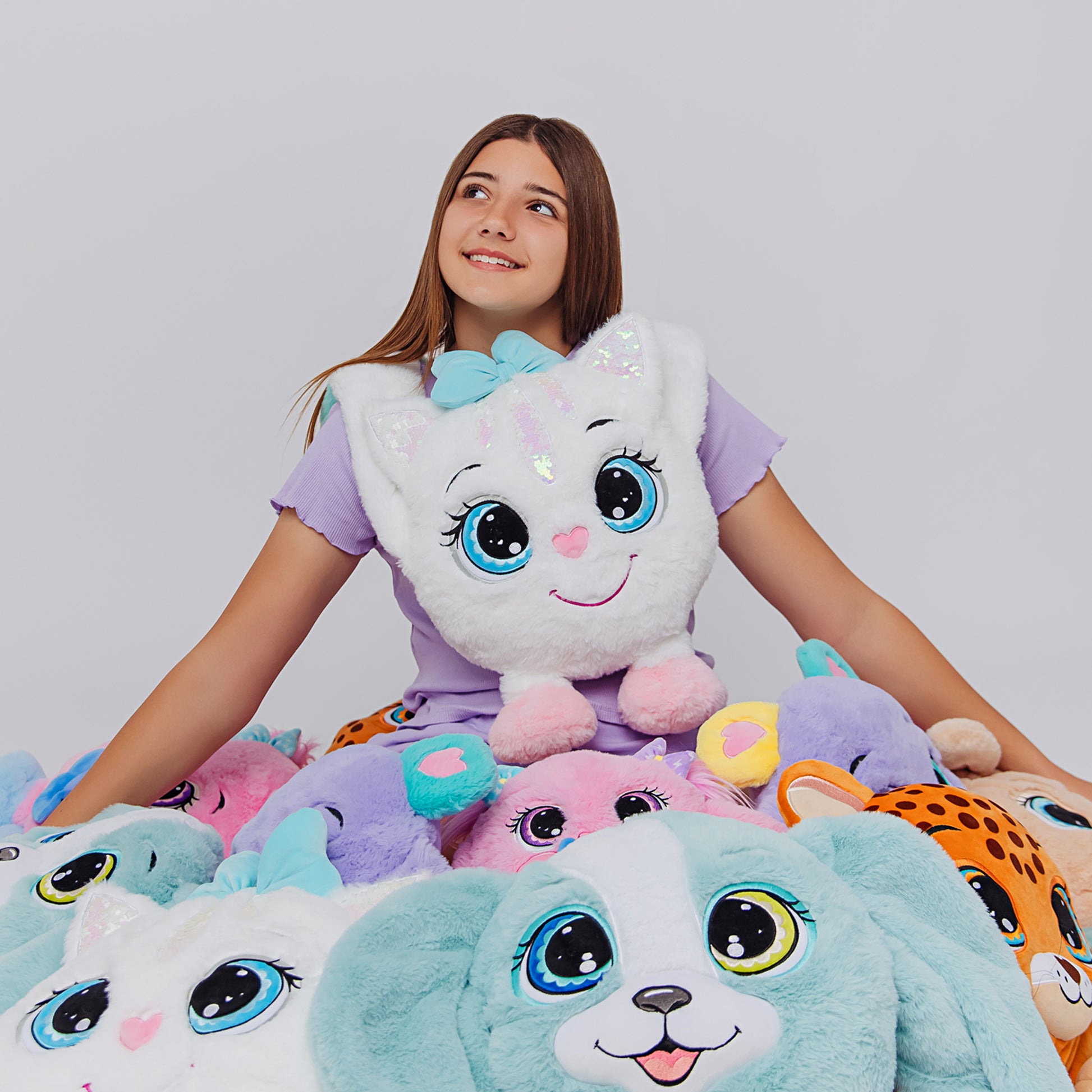 Girl holding a large weighted plush toy with a white face and blue accents, surrounded by similar plush toys on a plain background.